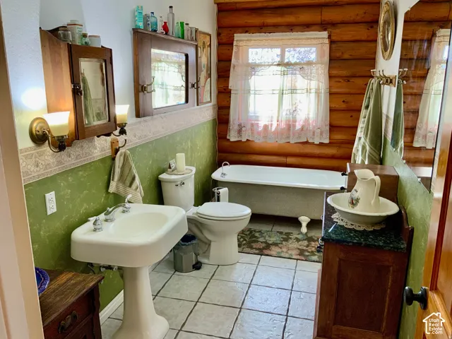 Pedestal sink with chrome fixtures, clawfoot tub, log wall paneling, tiled flooring, and wood-finish cabinetry