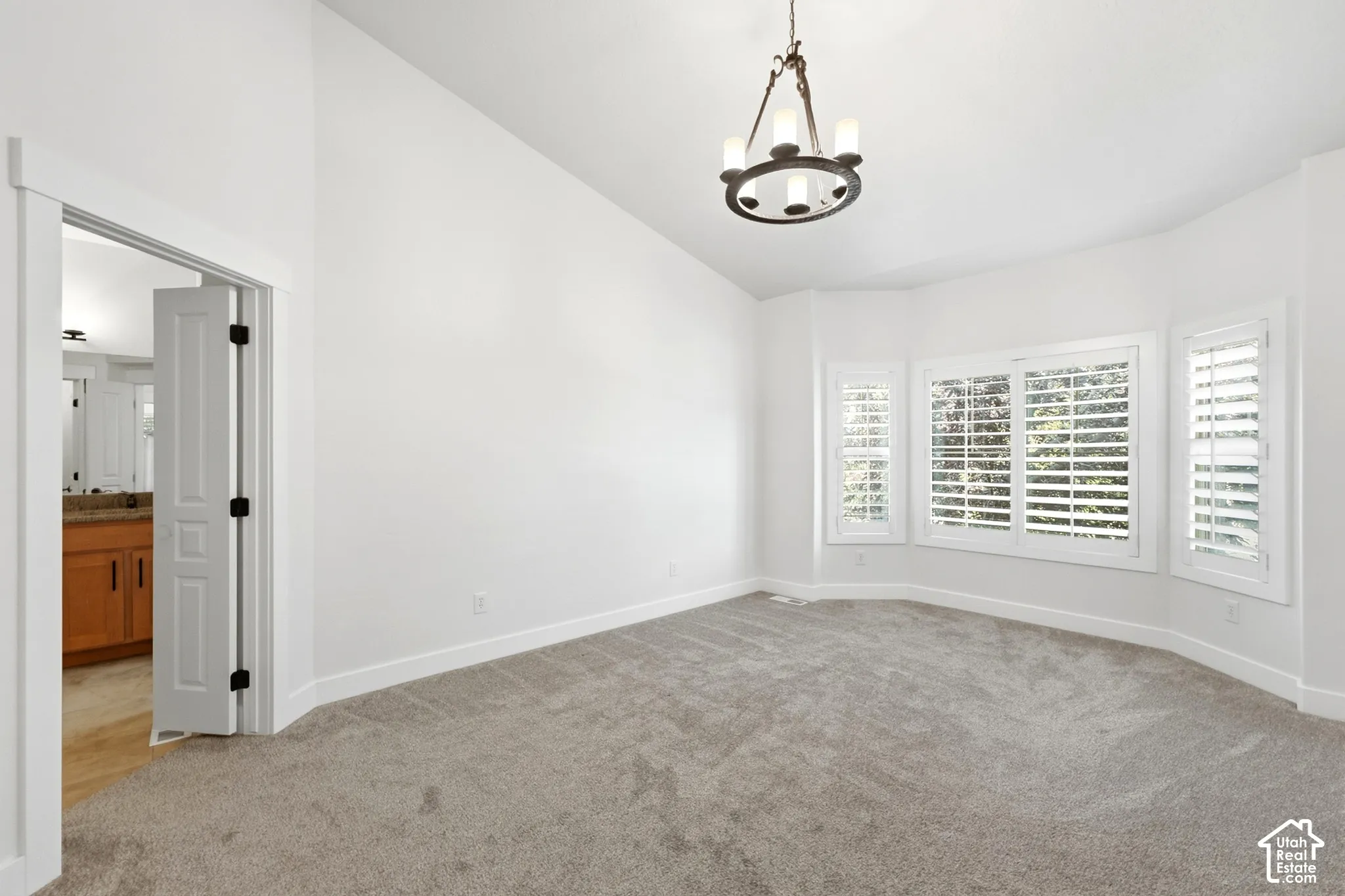 Vaulted ceiling, bay window with plantation shutters, ceiling-mounted chandelier, and grey carpeting