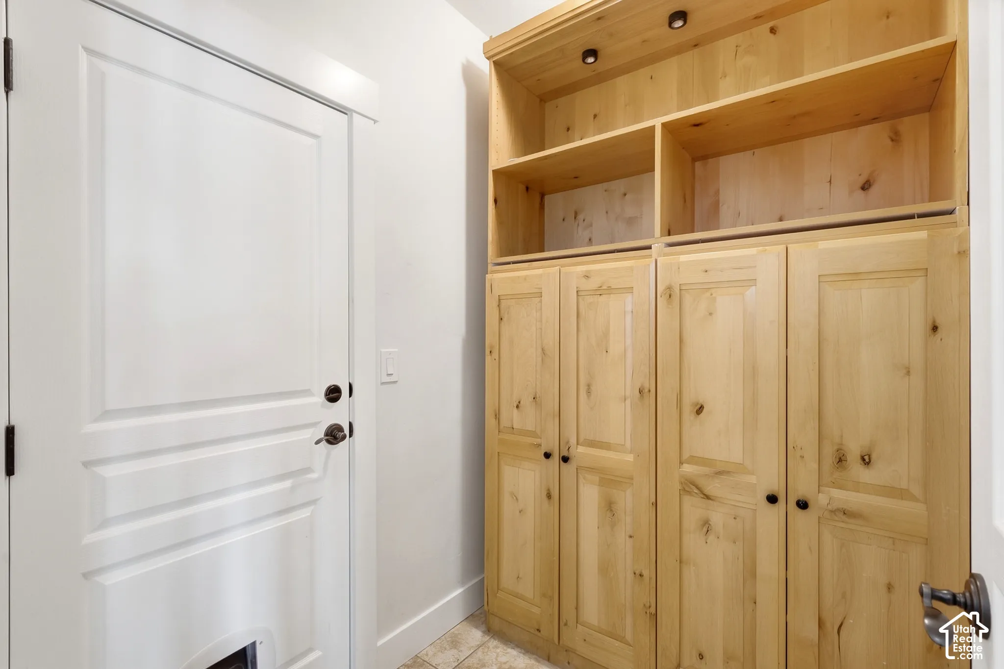 White paneled door with dark hardware, natural wood-finish built-in cabinetry, upper open shelving, recessed lighting, and light-toned tile flooring