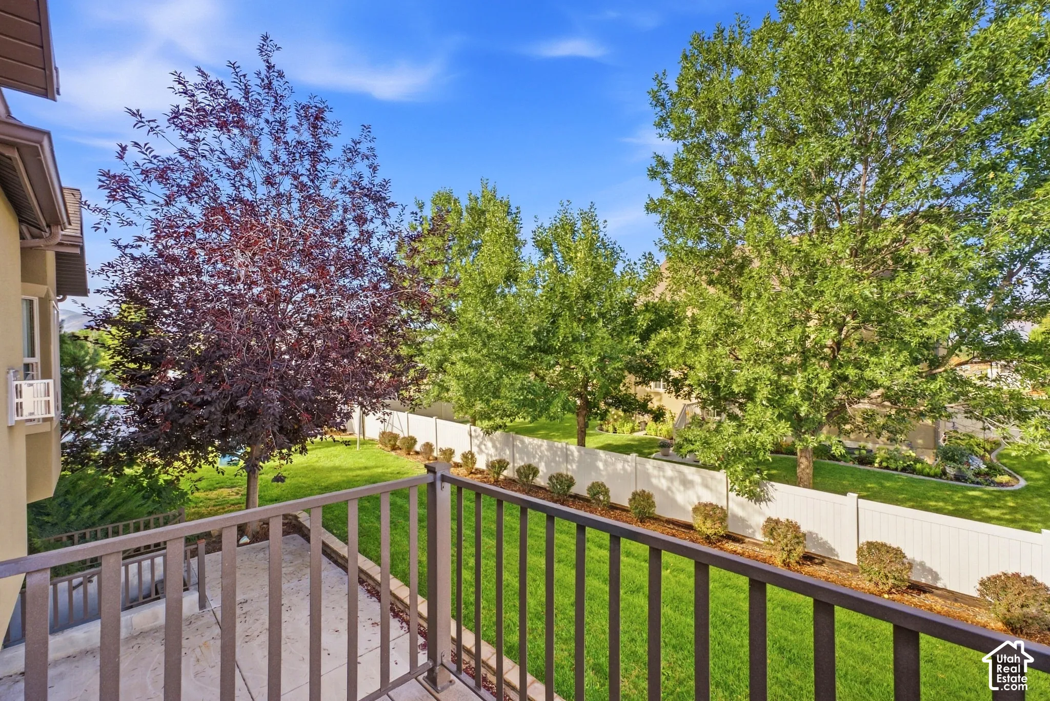 Balcony featuring metal railings and a concrete surface, overlooking a manicured lawn with mature trees and a privacy fence