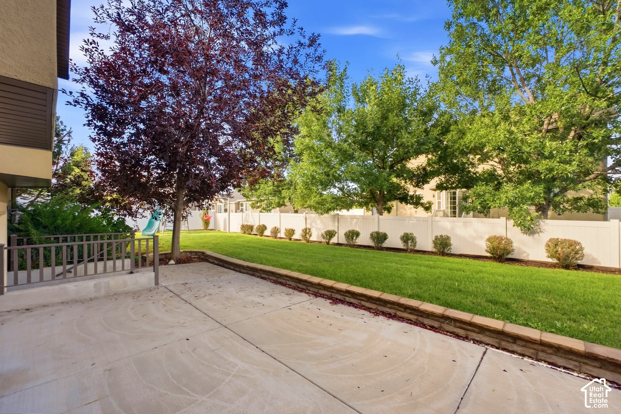Paved patio area, mature trees, manicured lawn, retaining wall, and a privacy fence