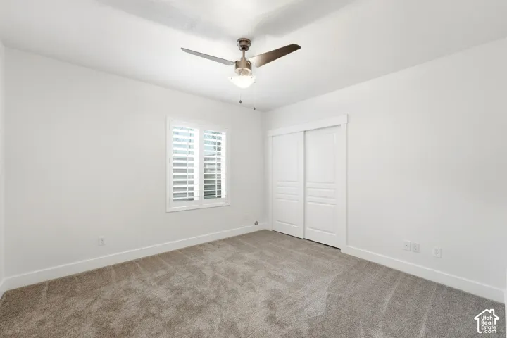 Neutral tone walls with white trim, gray carpet flooring, window with white plantation shutters, and a ceiling fan with light fixture