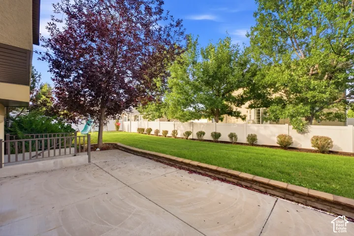 Paved patio area, mature trees, manicured lawn, retaining wall, and a privacy fence