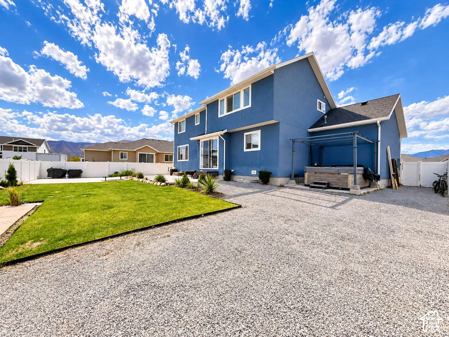 Two-story stucco exterior, expansive gravel patio, attached pergola, manicured lawn, and white vinyl fencing