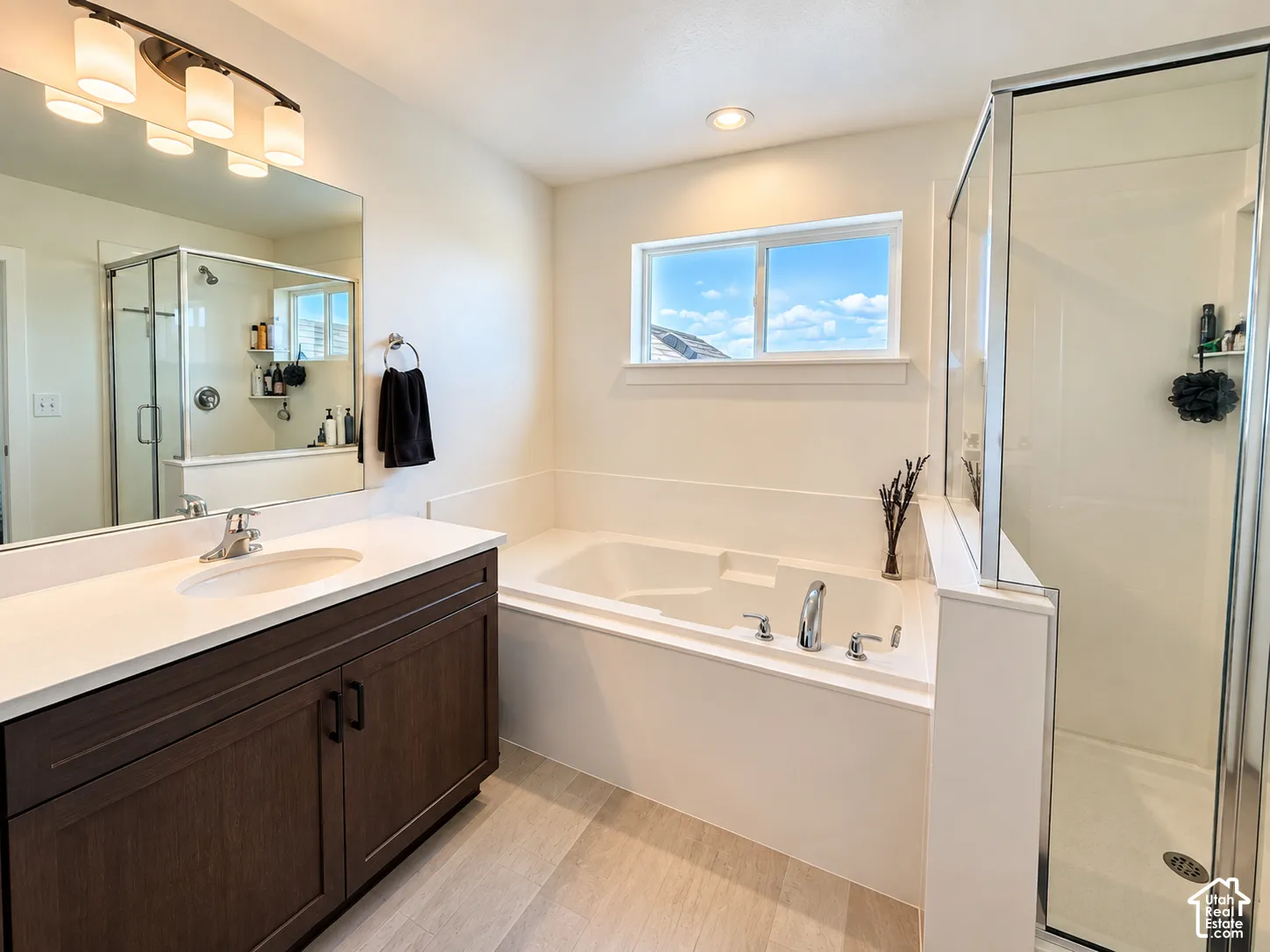 Wood-finish flooring, built-in soaking tub, glass-enclosed shower, single vanity with quartz countertop, and a large mirror