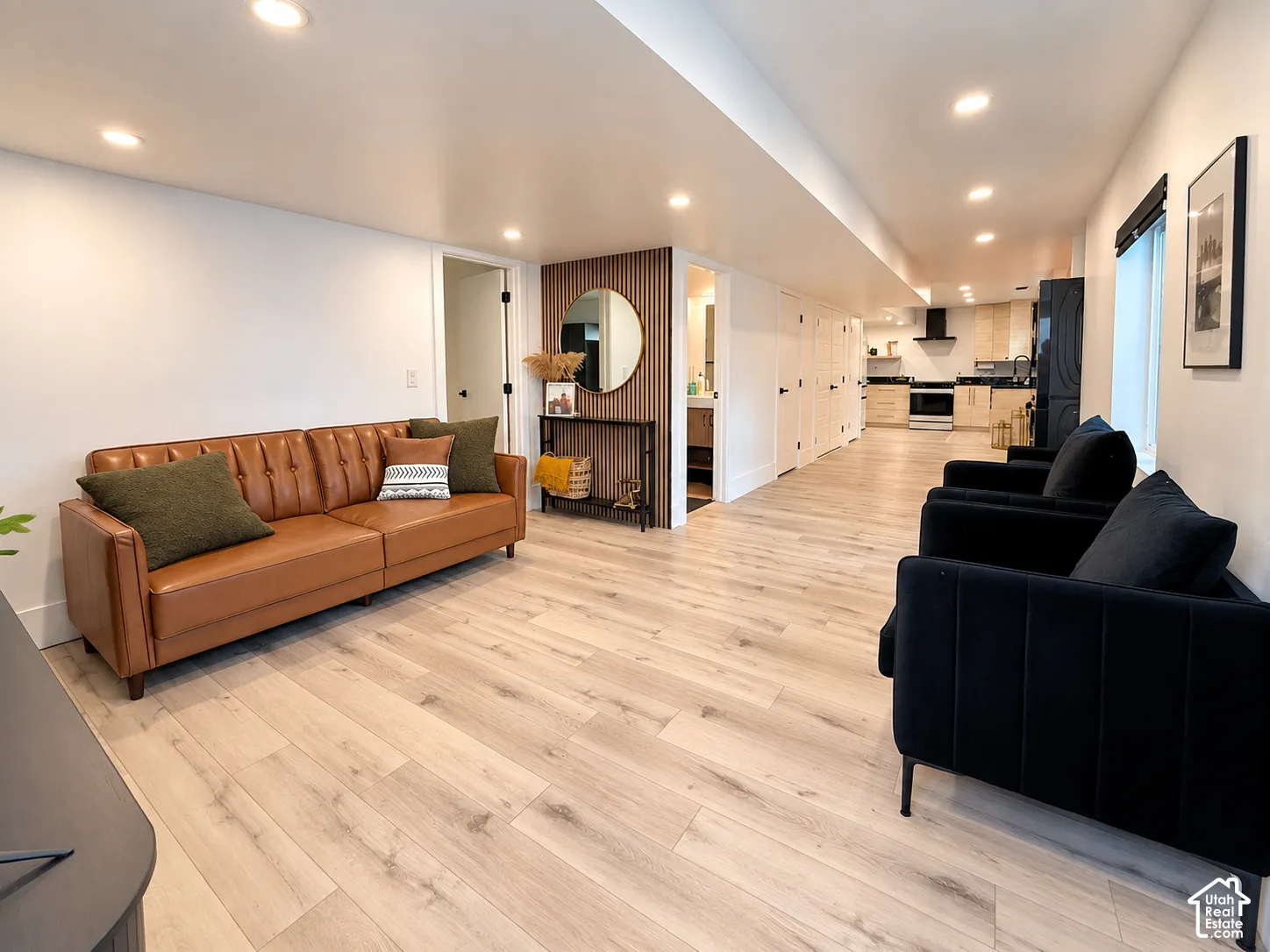 Recessed lighting illuminates the open-concept living space with wood-finish flooring, white walls, and a visible kitchen featuring light-toned cabinetry and a black range hood
