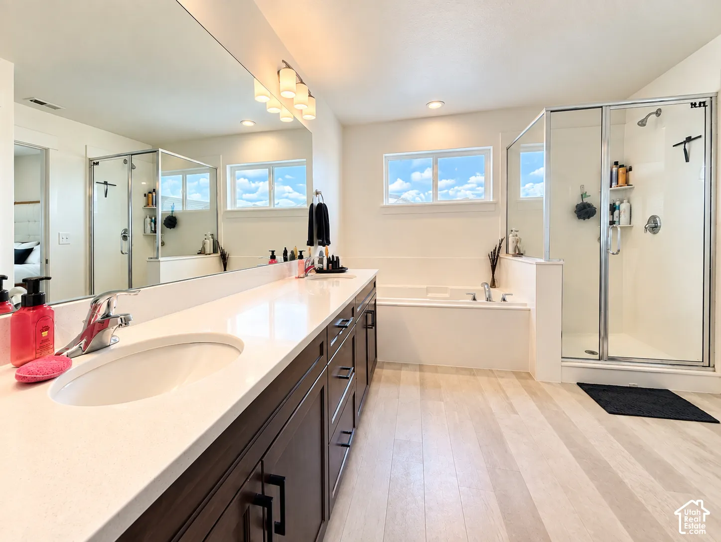 Spacious bathroom with light wood-finish flooring, a double vanity featuring a light-toned countertop and dark cabinetry, a built-in bathtub, and a glass-enclosed shower
