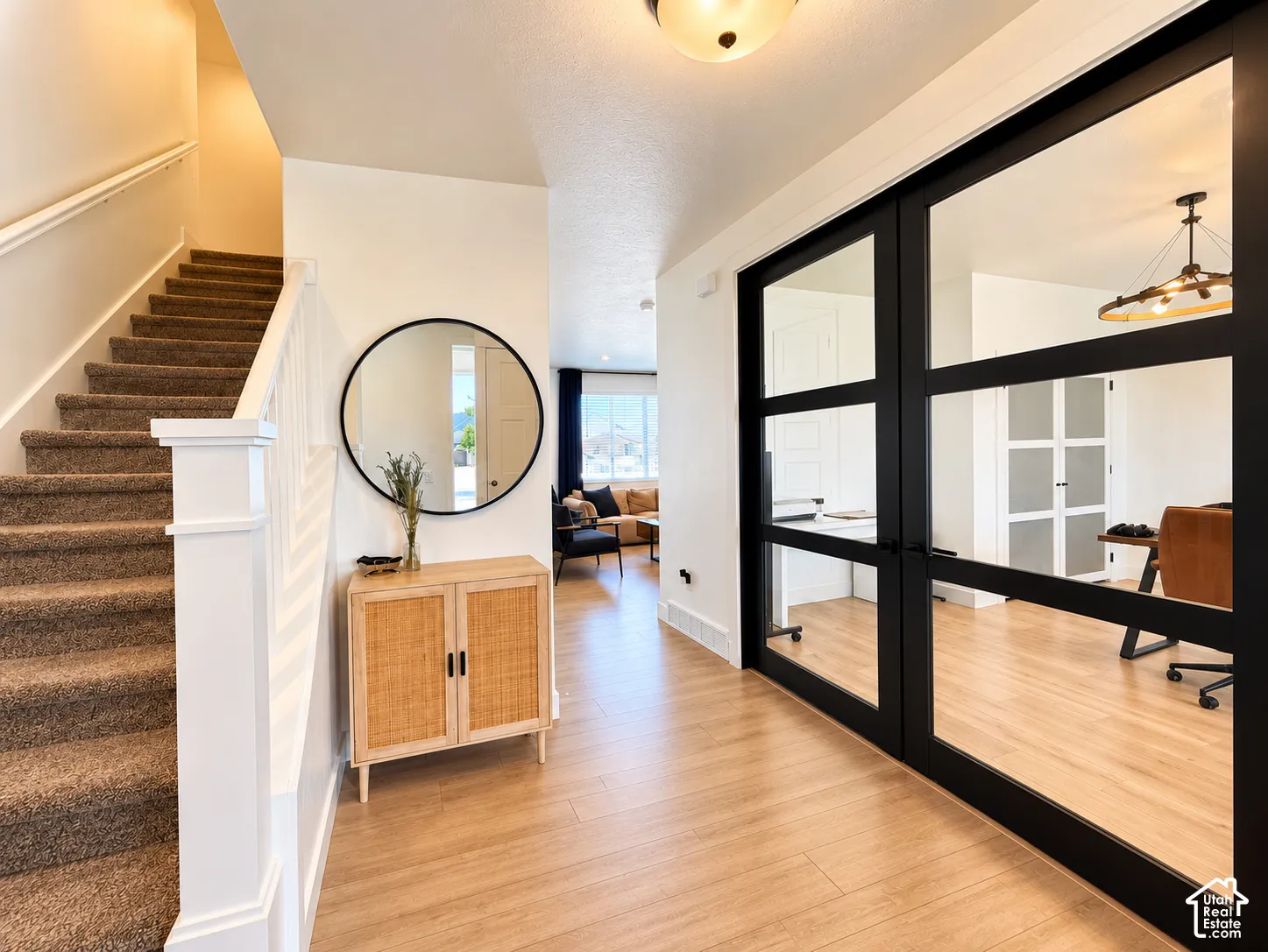 Wood-finish flooring, staircase with carpeted treads and white banister, black-framed glass double doors, and an overhead flush-mount light fixture