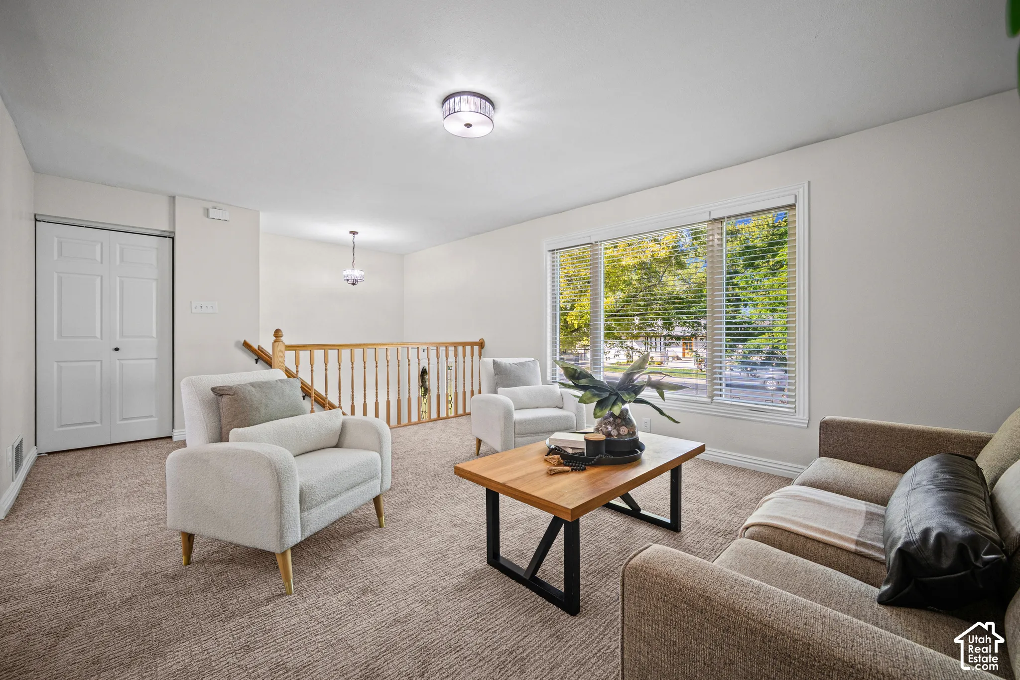 Bright and spacious living area showcasing neutral carpeting, large windows with natural light, curved upholstered seating, a wood-top coffee table with metal frame base, recessed ceiling lighting, and direct access to an adjacent staircase with wooden ra