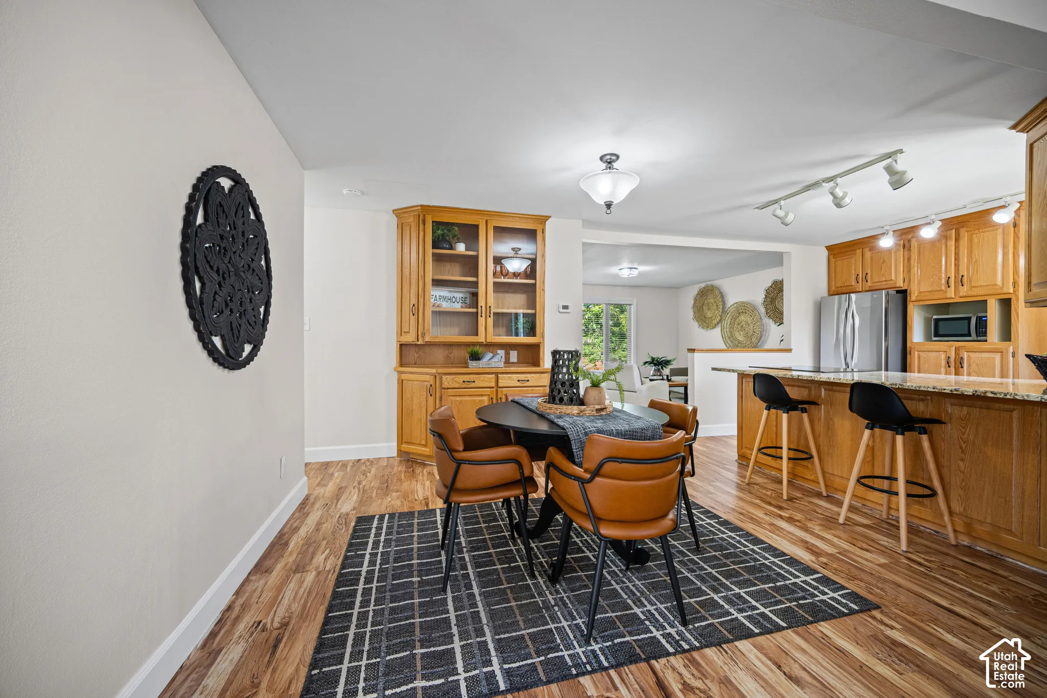 Open-concept dining and kitchen area featuring wood cabinetry with glass-front upper storage, stainless steel appliances, wood-finish flooring throughout, and a geometric patterned area rug anchoring the dining space with mid-century modern seating