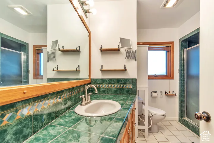 Bathroom featuring a single basin vanity with a tile countertop, a framed mirror, a window with wood trim, a toilet, and a glass-enclosed shower