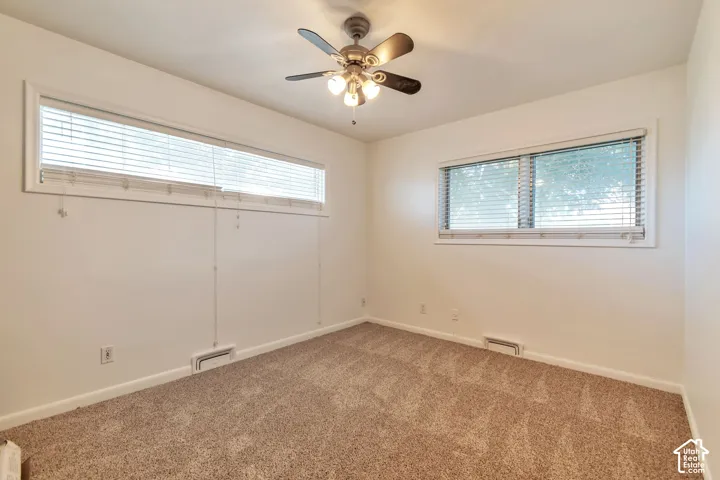 Neutral wall color with plush carpet flooring, two windows with horizontal blinds, and a ceiling fan with integrated lighting