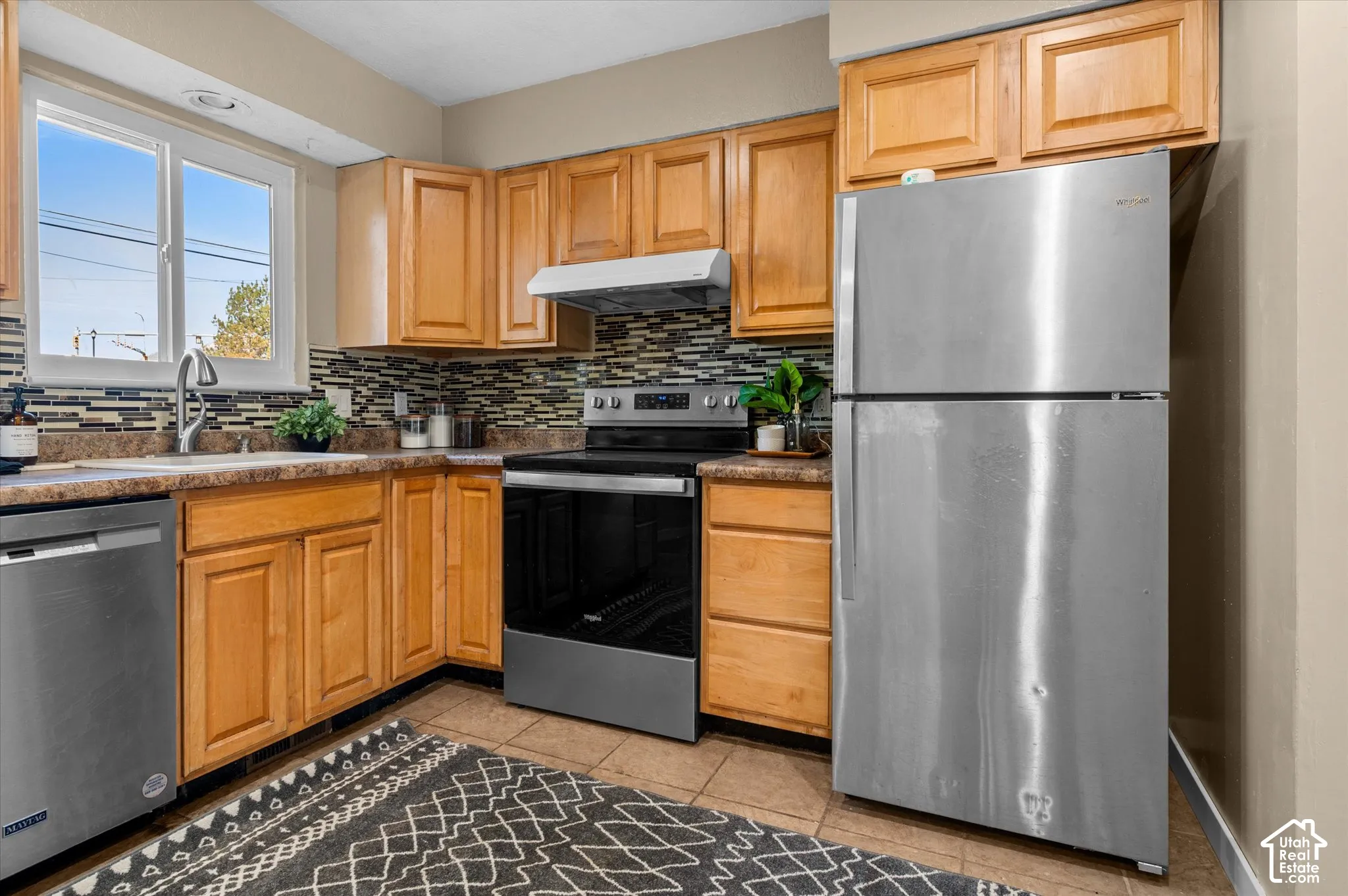 Wood-finish cabinetry, stainless steel appliances, mosaic tile backsplash, and a window above the sink