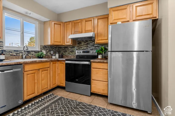 Wood-finish cabinetry, stainless steel appliances, mosaic tile backsplash, and a window above the sink