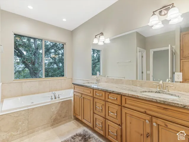 Built-in soaking tub, large window, dual vanity with undermount sinks, and light wood cabinetry with stone countertops
