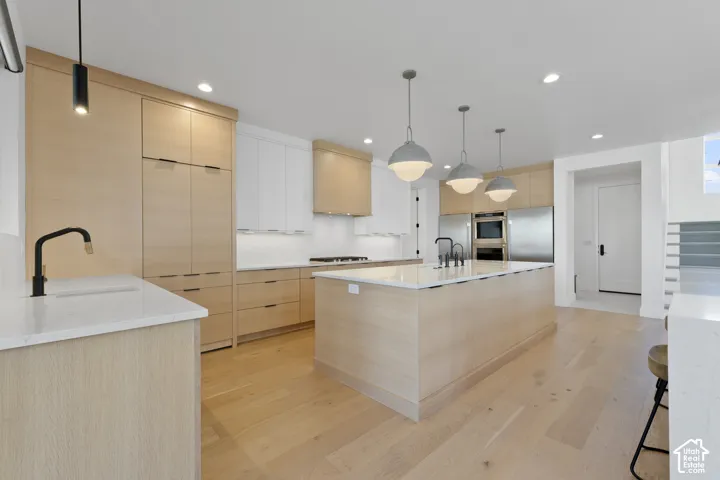 Modern kitchen featuring light wood-finish flooring, light wood cabinetry, white upper cabinetry, stainless steel appliances, and a central island with pendant lighting