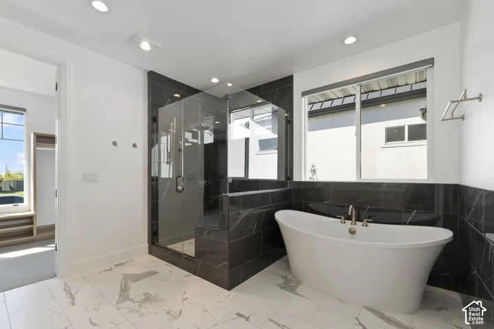 Freestanding soaking tub with a floor-mounted faucet, walk-in shower with glass enclosure, dark-toned tile surround, and white marble-look flooring