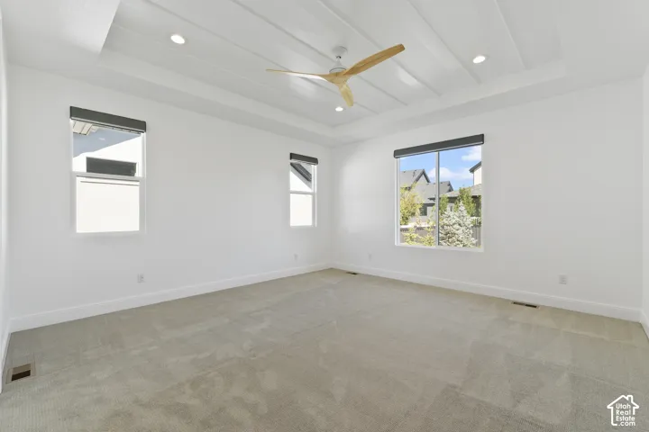 Tray ceiling with recessed lighting, large windows with roller shades, light-toned carpeting, and a ceiling fan