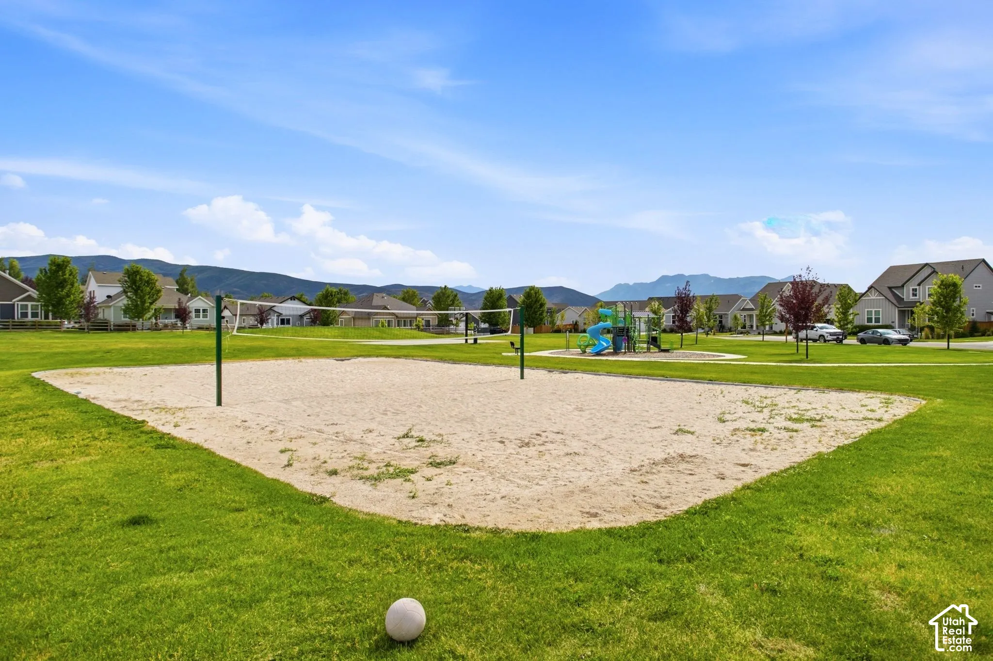 Expansive green space featuring a sand volleyball court, playground structure, and mature trees