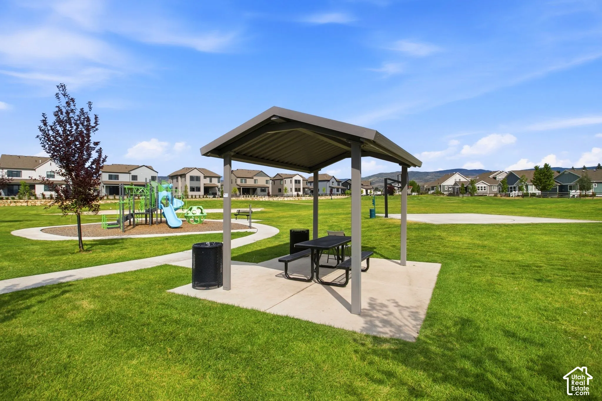 Community park featuring a spacious open lawn, covered picnic shelter with integrated seating and dining surface, concrete pads, trash receptacle, and mature landscaping with established trees throughout the grounds