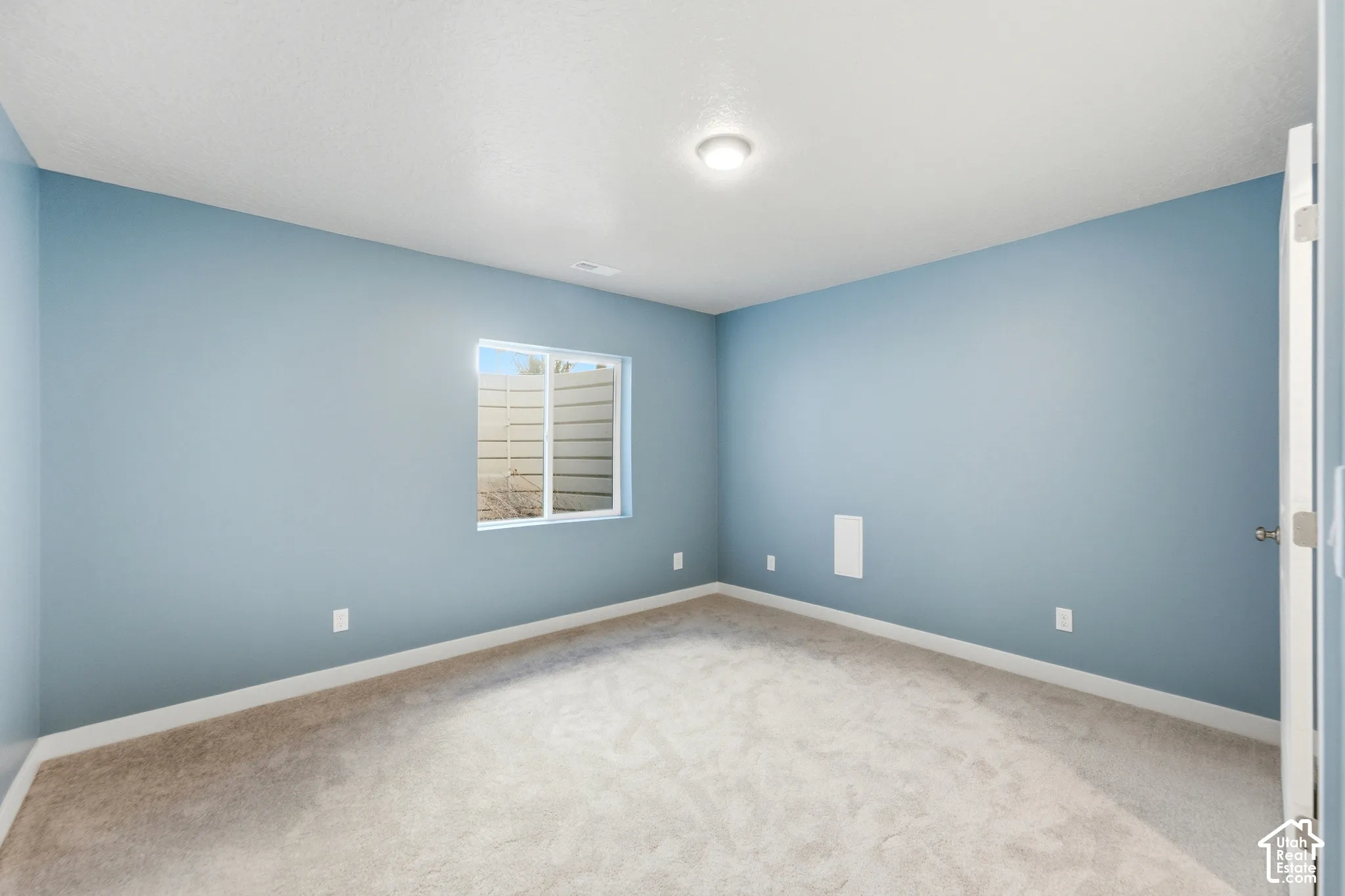 Spacious bedroom showcasing soft blue wall finishes, neutral carpet flooring, recessed ceiling lighting, a large window with plantation shutters providing natural light, and multiple electrical outlets throughout