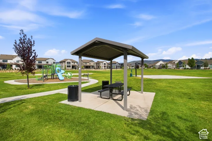 Community park featuring a spacious open lawn, covered picnic shelter with integrated seating and dining surface, concrete pads, trash receptacle, and mature landscaping with established trees throughout the grounds