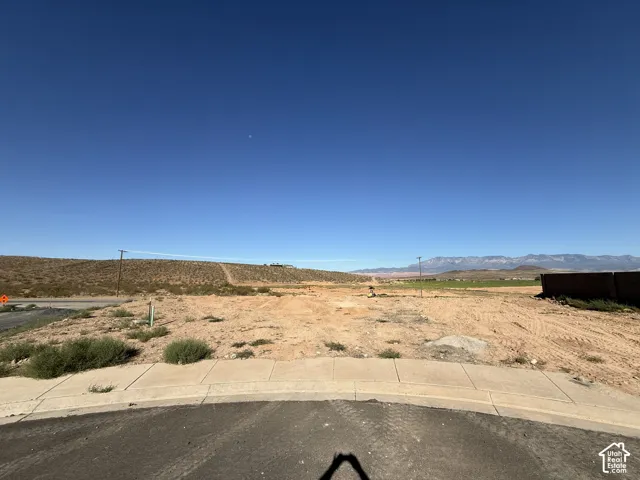 Paved road frontage with a concrete sidewalk, open land, and distant mountains