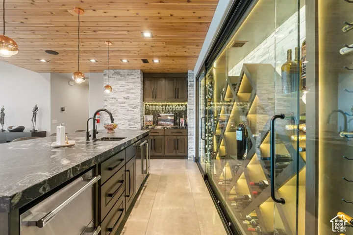 Expansive wine cellar with custom shelving and glass doors, island with dark stone countertop, built-in sink with black faucet, wood-finish ceiling, and recessed lighting