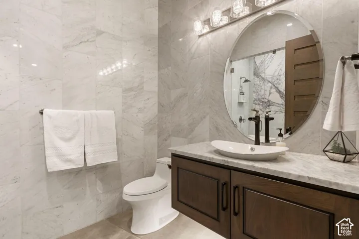 Full wall tiling, wood-finish cabinetry, countertop with a vessel sink, round wall mirror, and a multi-bulb vanity light fixture
