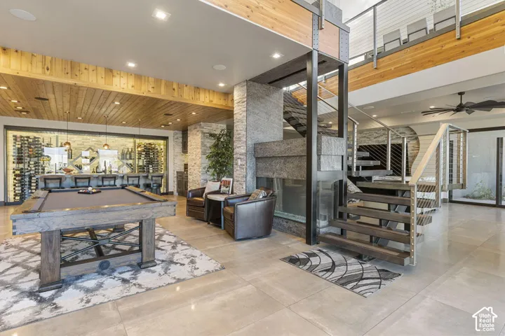 Open-concept interior featuring light-toned tile flooring, a built-in bar with extensive wine storage, a contemporary staircase with metal railings, a stone-clad column, and a wood-paneled ceiling