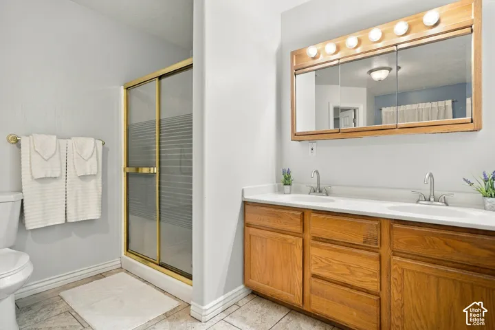 Dual basin vanity with wood-finish cabinetry and white countertop, framed mirror with integrated lighting, and a shower enclosure with gold-finish trim