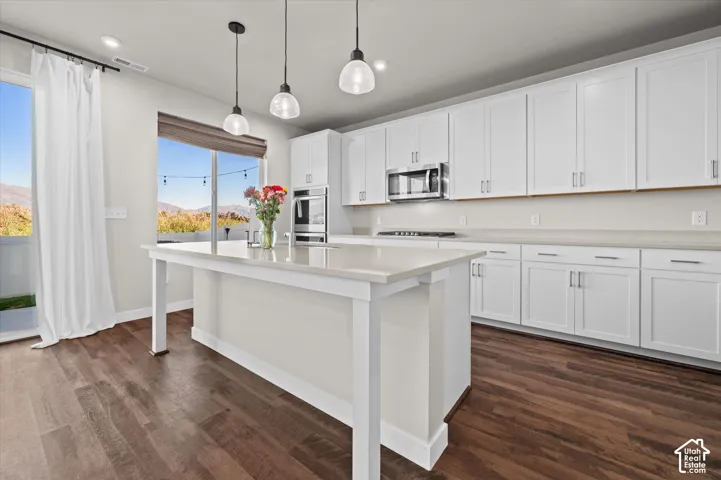 Kitchen featuring white shaker cabinetry, stainless steel appliances, a large center island with a light-toned countertop, wood-finish flooring, and pendant lighting