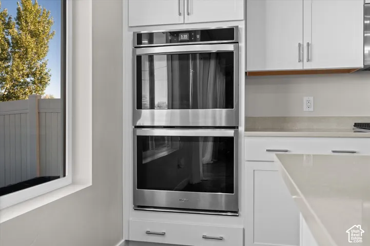 Stainless steel double wall ovens, white cabinetry with silver hardware, light-toned countertop, and a window with a white frame