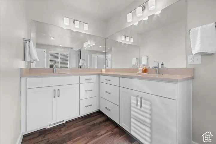 Corner vanity with dual sinks, light-toned countertops, white cabinetry, wood-finish flooring, and expansive wall mirrors