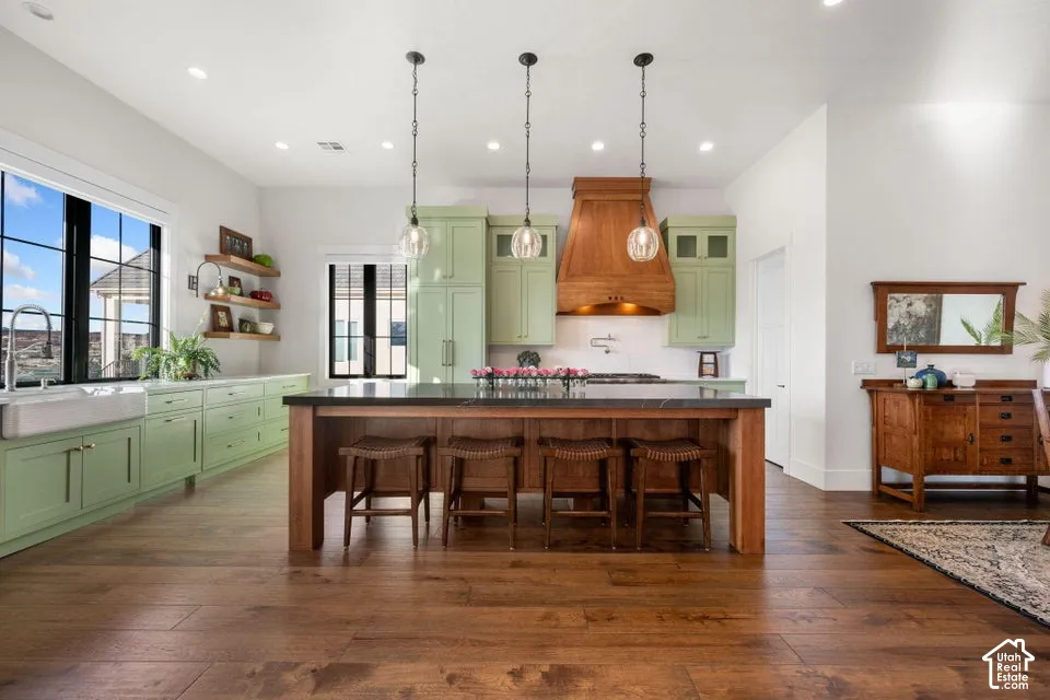 Wood-finish flooring, light green cabinetry, a large kitchen island with seating, a farmhouse sink, and a wood-panel range hood