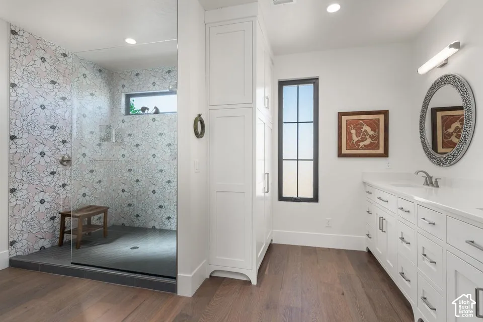 Glass-enclosed shower with decorative tiled walls, expansive vanity with dual sinks, built-in storage cabinetry, wood-finish flooring, and a large window
