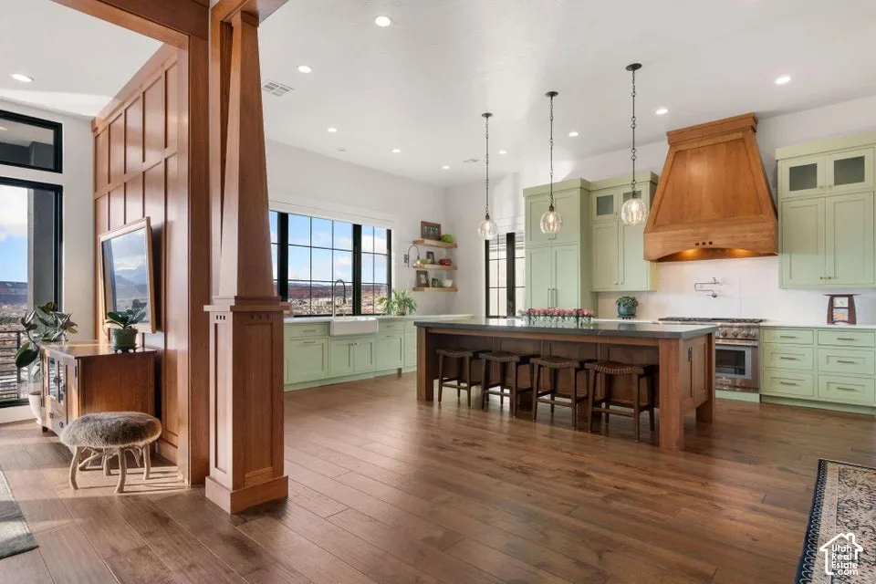Hardwood-finish flooring extending throughout, a kitchen island with seating, pendant lighting, custom wood range hood, and light green cabinetry