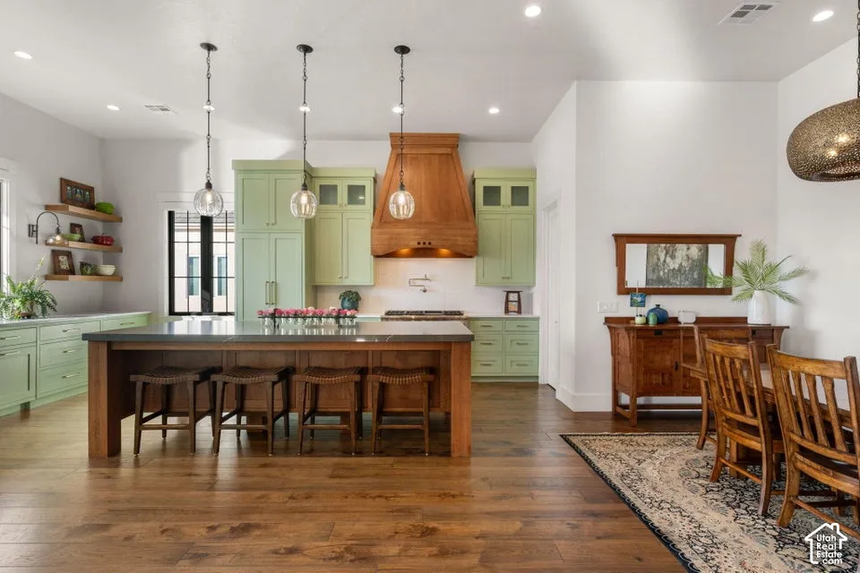 Open concept kitchen featuring an oversized island with seating, pendant lighting, custom wood vent hood, green cabinetry, and wood-finish flooring