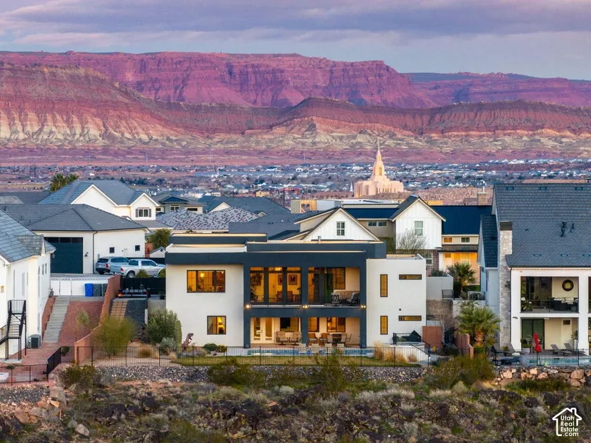 Modern architecture with clean-lined exterior featuring white and dark composite siding, expansive multi-pane glass doors and windows with warm interior lighting, pitched roof with slate-colored shingles, and manicured desert landscaping with native plant