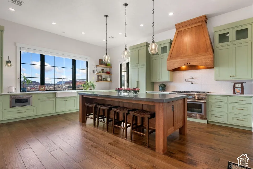 Kitchen featuring wood-finish flooring, light green cabinetry, a large center island with seating, a prominent wood range hood, and a built-in microwave