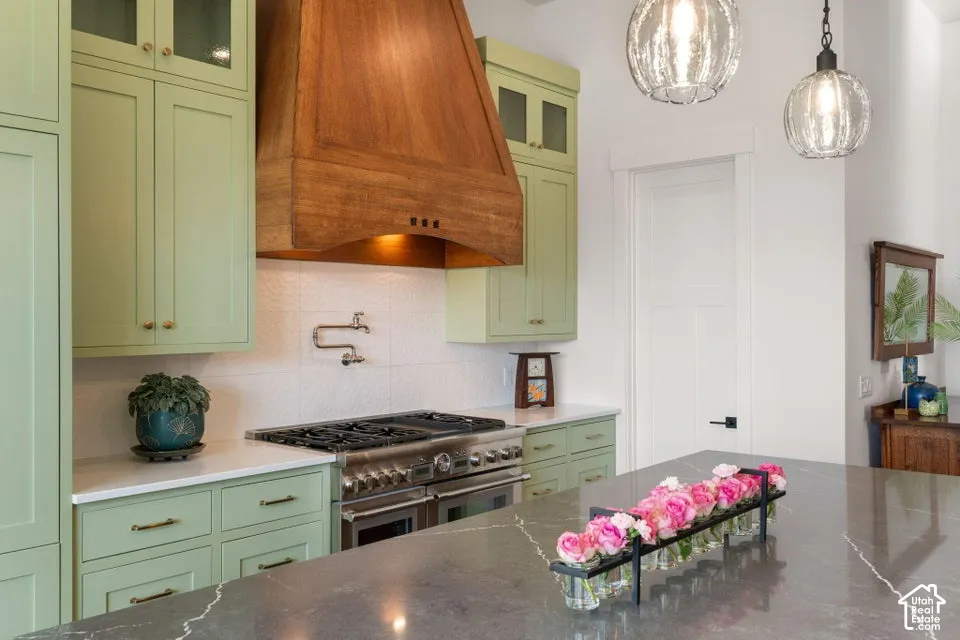 Sage green cabinetry with brass hardware, wood-finish range hood, stainless steel range, white textured backsplash, and a dark stone countertop island