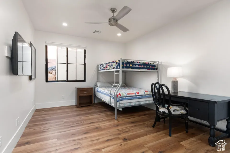 Bright secondary bedroom showcasing hardwood flooring, a metal bunk bed frame, black desk with seating, and a ceiling fan