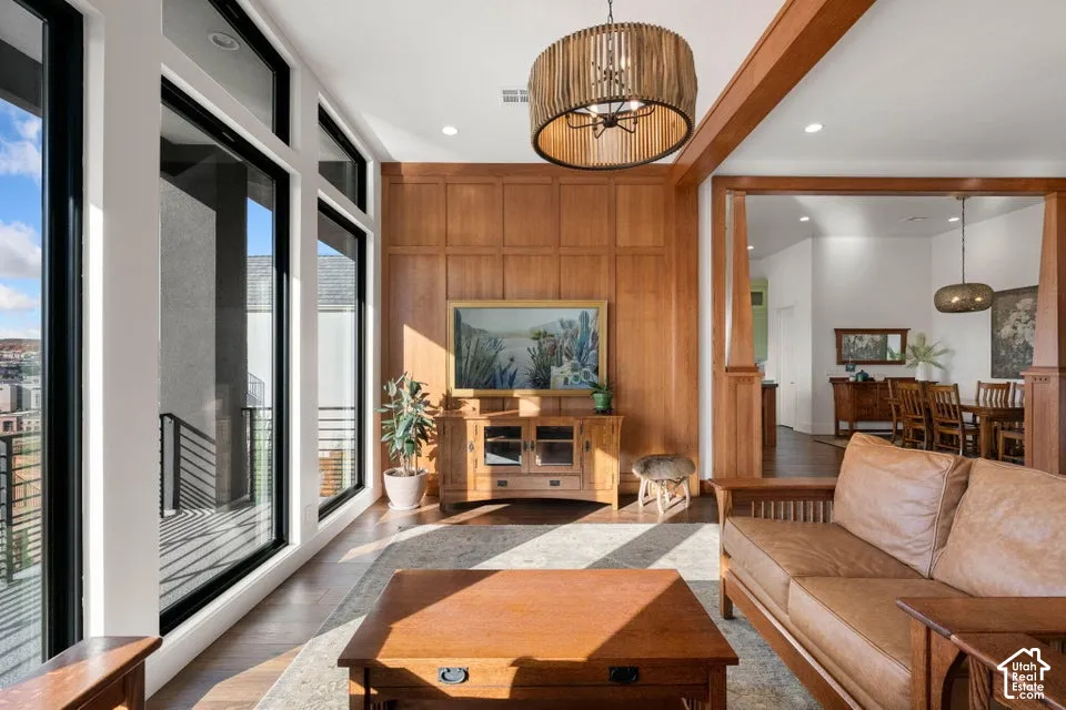 Expansive windows with black framing, wood-finish flooring, wood paneled wall, decorative ceiling light fixture, and an open entryway to an adjacent dining area