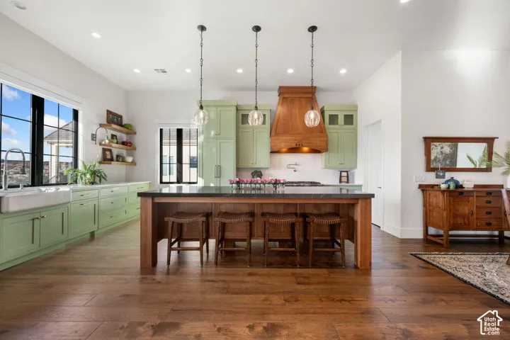 Wood-finish flooring, light green cabinetry, a large kitchen island with seating, a farmhouse sink, and a wood-panel range hood