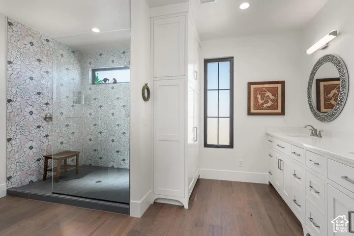 Glass-enclosed shower with decorative tiled walls, expansive vanity with dual sinks, built-in storage cabinetry, wood-finish flooring, and a large window