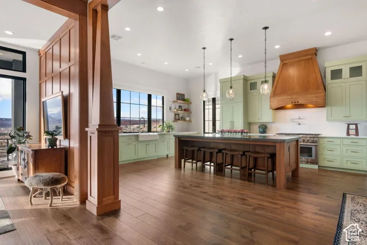 Hardwood-finish flooring extending throughout, a kitchen island with seating, pendant lighting, custom wood range hood, and light green cabinetry