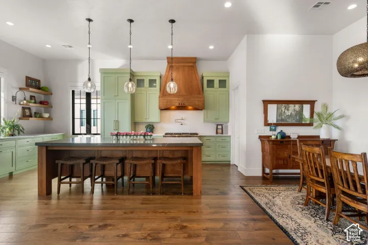 Open concept kitchen featuring an oversized island with seating, pendant lighting, custom wood vent hood, green cabinetry, and wood-finish flooring