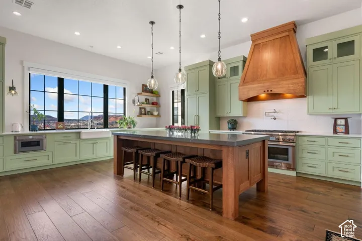 Kitchen featuring wood-finish flooring, light green cabinetry, a large center island with seating, a prominent wood range hood, and a built-in microwave