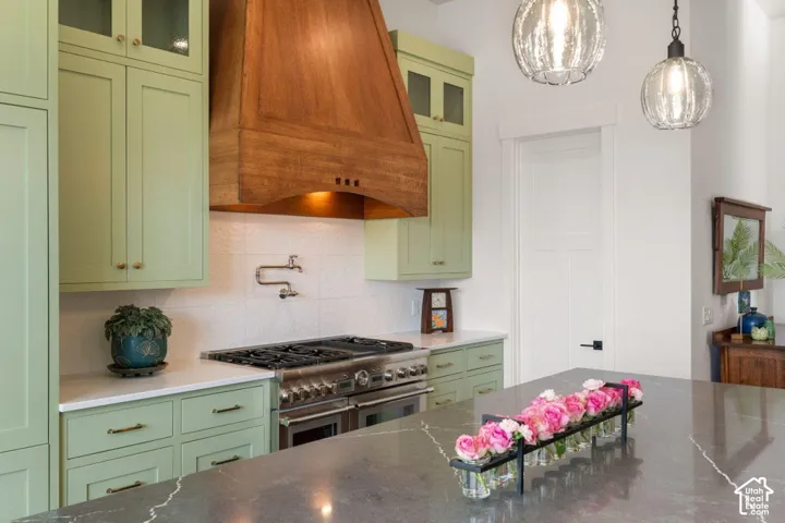 Sage green cabinetry with brass hardware, wood-finish range hood, stainless steel range, white textured backsplash, and a dark stone countertop island