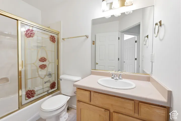 White bathtub with a gold-framed sliding glass enclosure, a white toilet, and a vanity with a light-toned countertop and wood-finish cabinetry